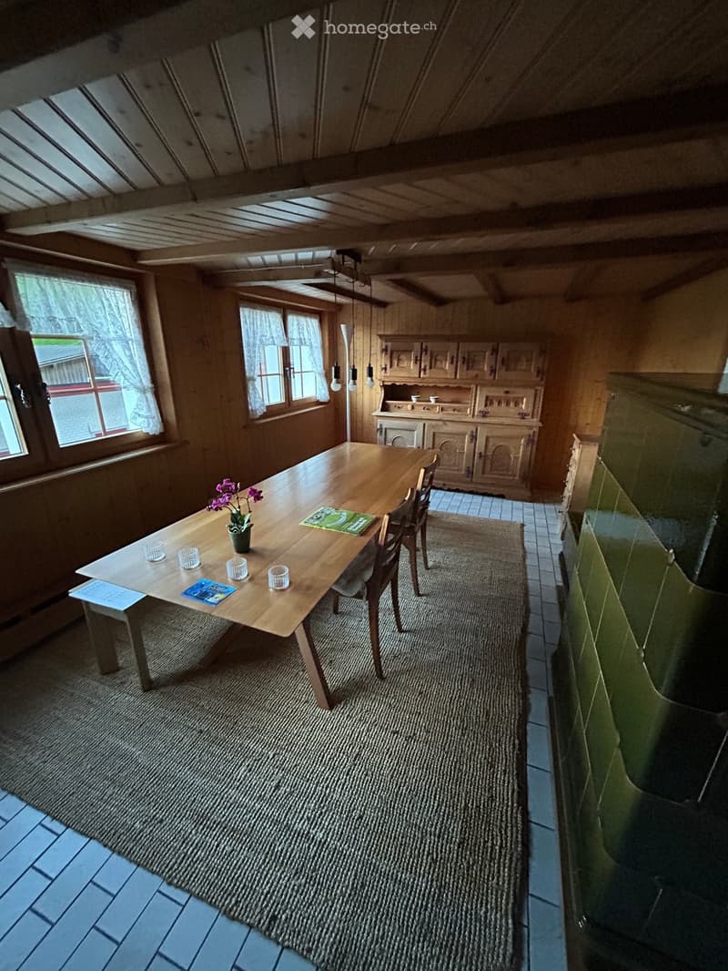 Cozy kitchen with wooden dining table, chairs, and a cabinet. The room features wooden walls, a tiled floor, and a large window with curtains.