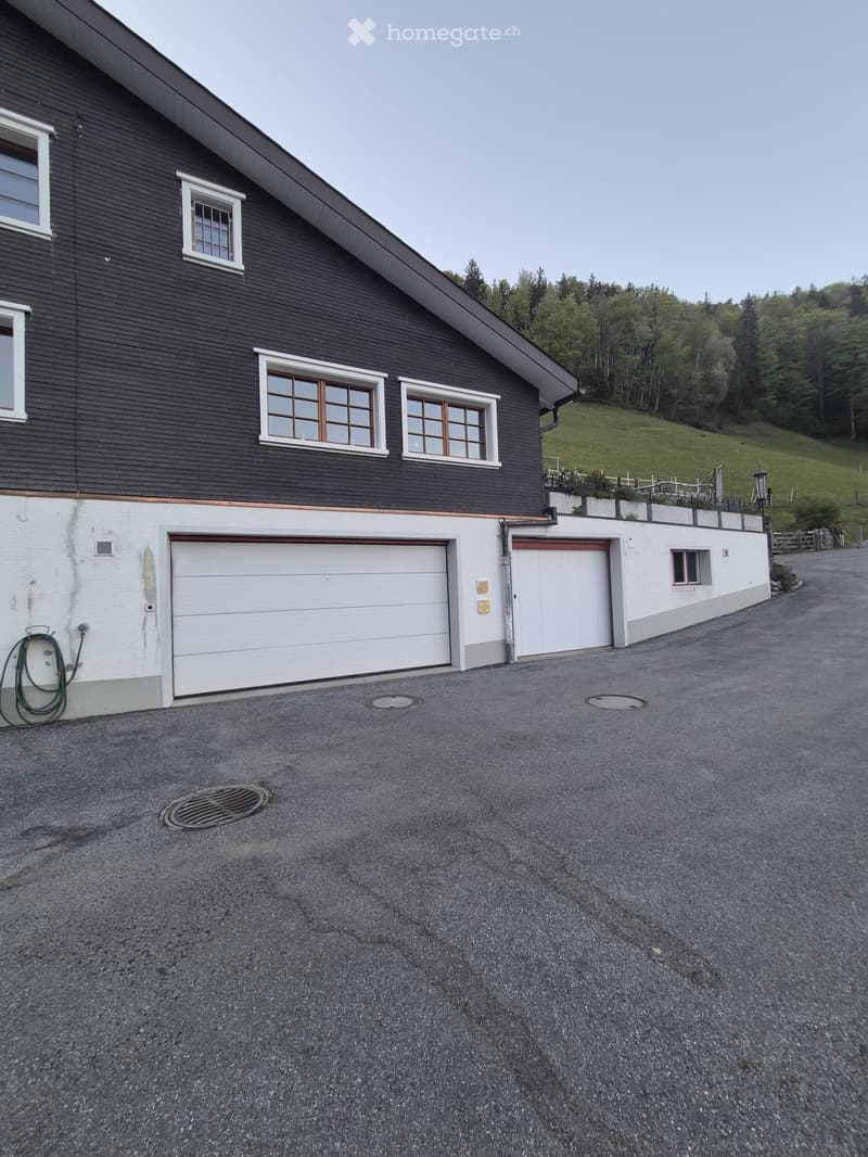 Two-story house with dark wooden siding, white garage doors, and multiple windows, set on a paved driveway with a green hillside in the background.