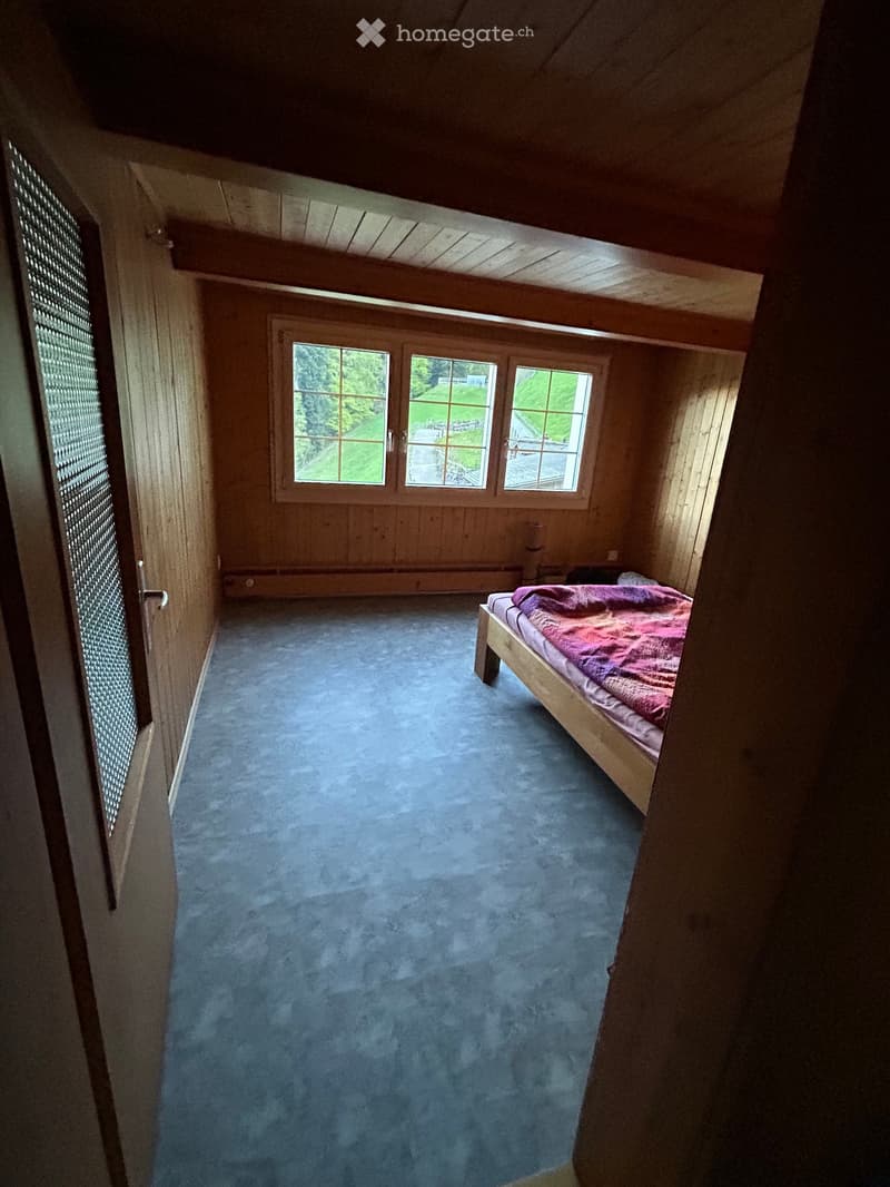 Cozy attic bedroom with wooden walls, slanted ceiling, blue carpet, and a single bed with a pink blanket.