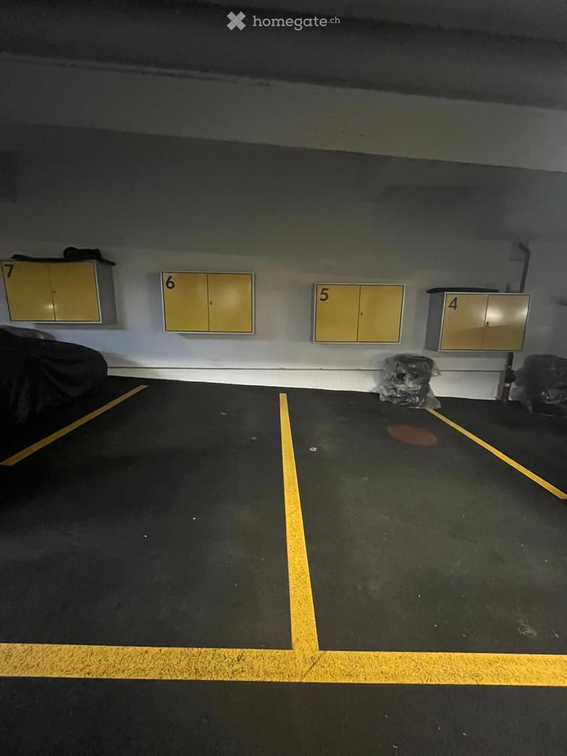 Underground parking area with numbered storage units, yellow lines marking parking spaces, and a covered vehicle partially visible.