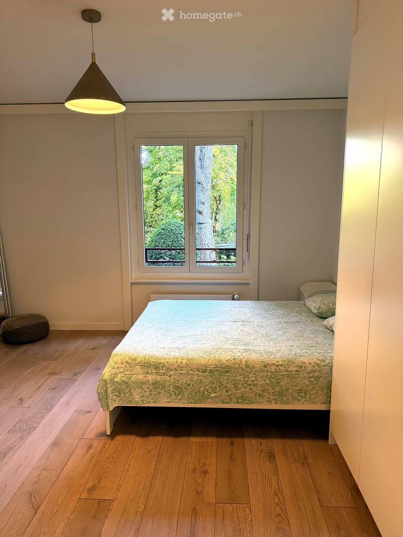 Minimalist bedroom with a single bed, wooden flooring, and a hanging pendant light. A small stool is placed near the bed, and a window offers a view of greenery outside.