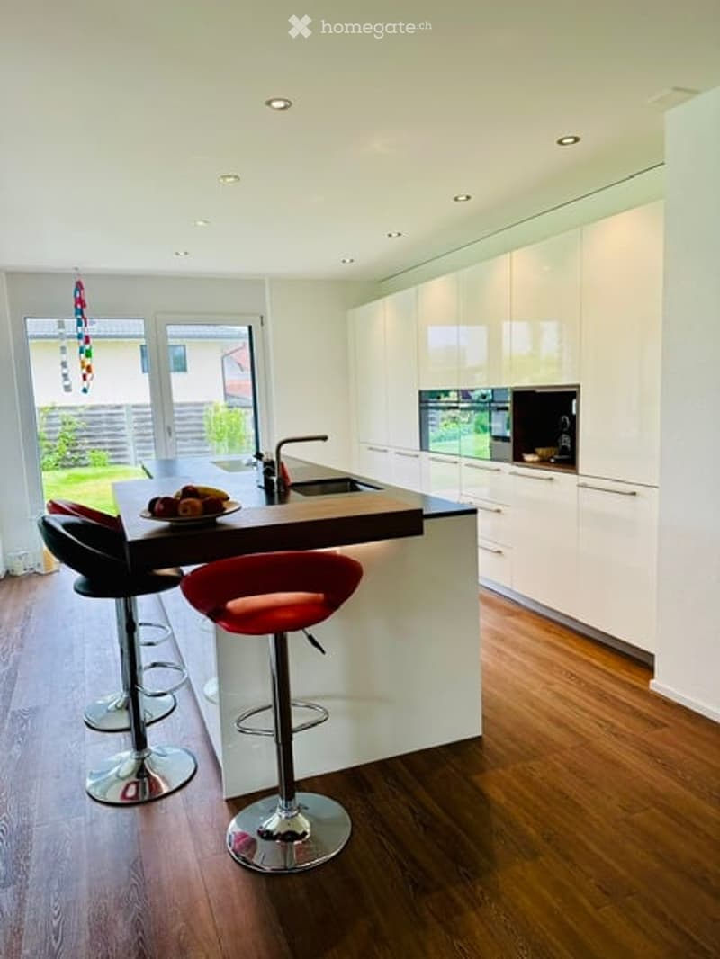 Modern kitchen with white cabinets, a central island, and red bar stools. Features a sink, oven, and a plate with fruit on the island.