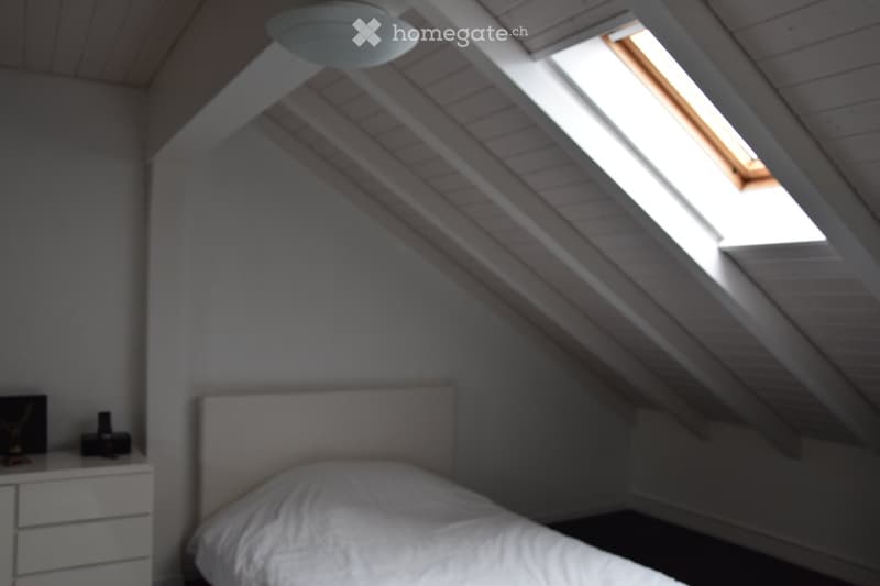 Cozy attic bedroom with slanted wooden ceilings, skylight window, white bed, nightstand, and lamp.