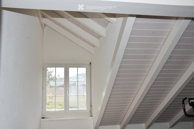 An attic space with wooden slatted ceilings, white walls, and a large window offering a view of the outdoors. The room is empty, with no furniture or furnishings visible.
