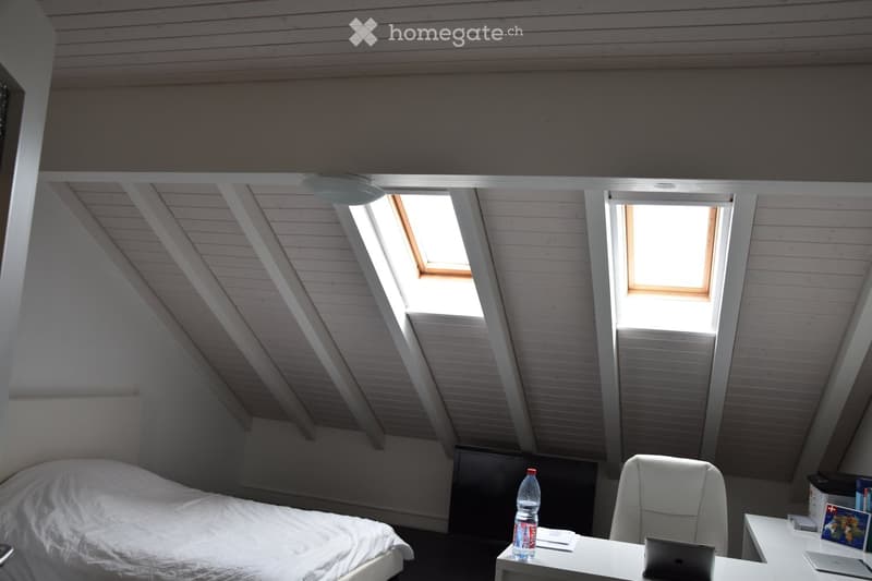Cozy attic bedroom with slanted wooden ceilings, skylights, a neatly made bed, a white desk with a laptop, and a chair.