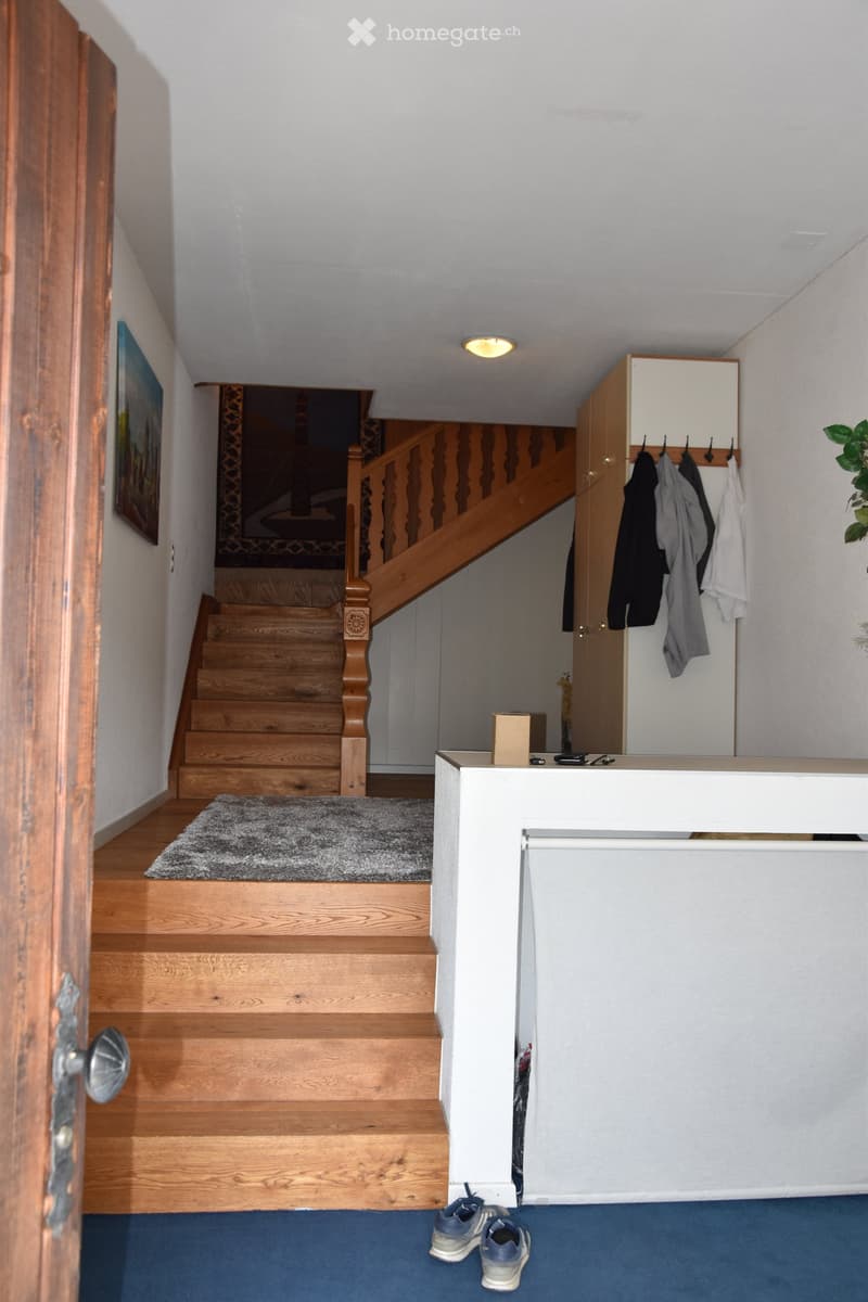 Hallway with wooden staircase, coat rack, and a small reception area. Blue carpet, white walls, and a painting on the wall.