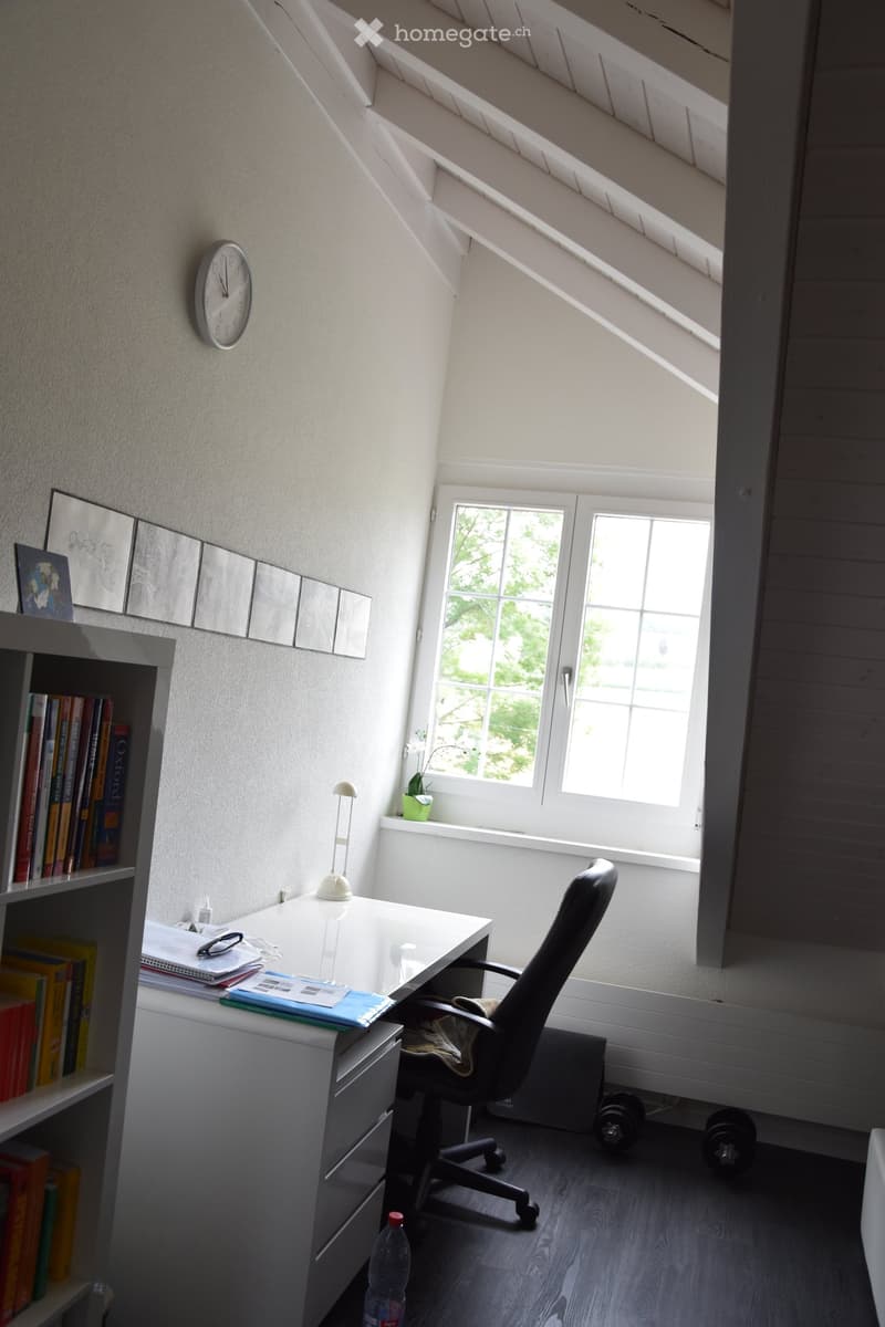 A cozy attic study with a slanted ceiling, large window, desk, chair, bookshelf, clock, and small plant on the windowsill.