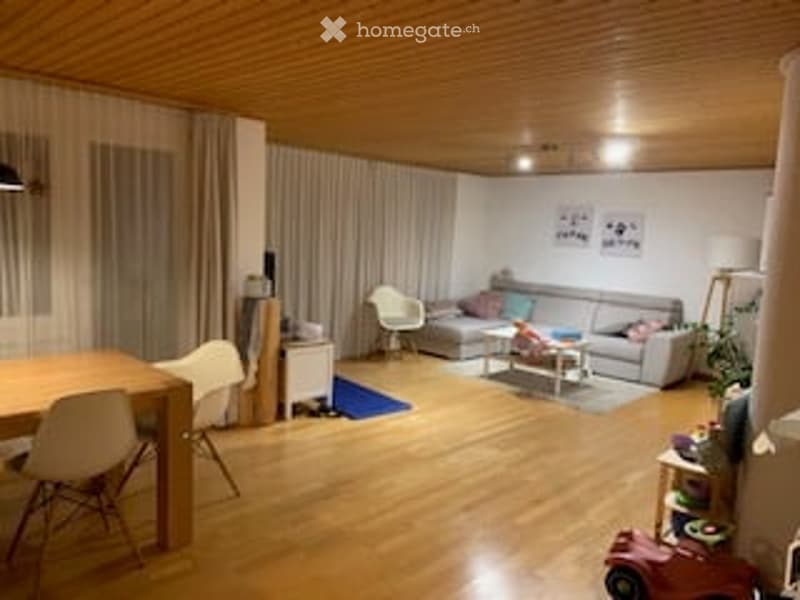 Bright, open-plan living and dining area with wooden flooring, large windows with curtains, grey sofa, coffee table, dining table with chairs, and decorative items.