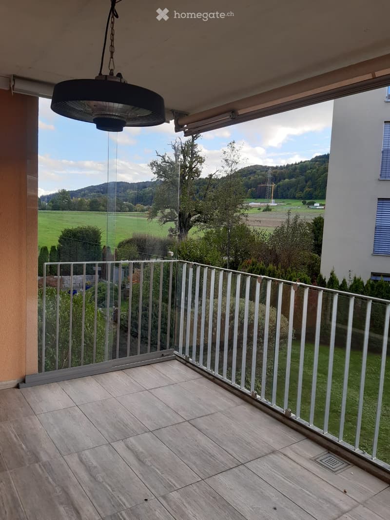 Balcony with tiled floor, glass railing, and a view of a green field and hills.