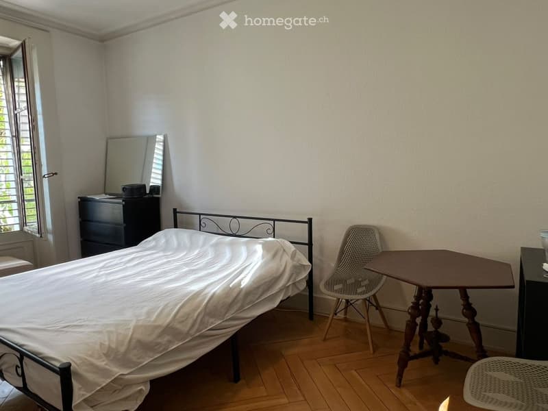 A small bedroom with a single bed, a wooden nightstand, a small table with a chair, and a mirror on the wall. The room has wooden flooring and a window with shutters.