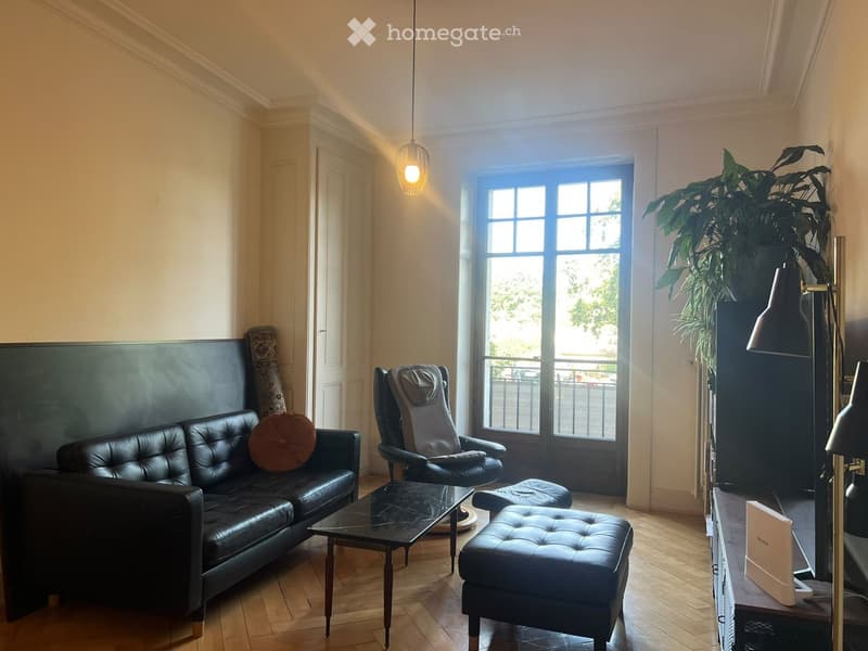 Modern living room with a black leather sofa, coffee table, ottoman, and a large plant near the window. The room features wooden flooring and large windows with a view of the balcony.