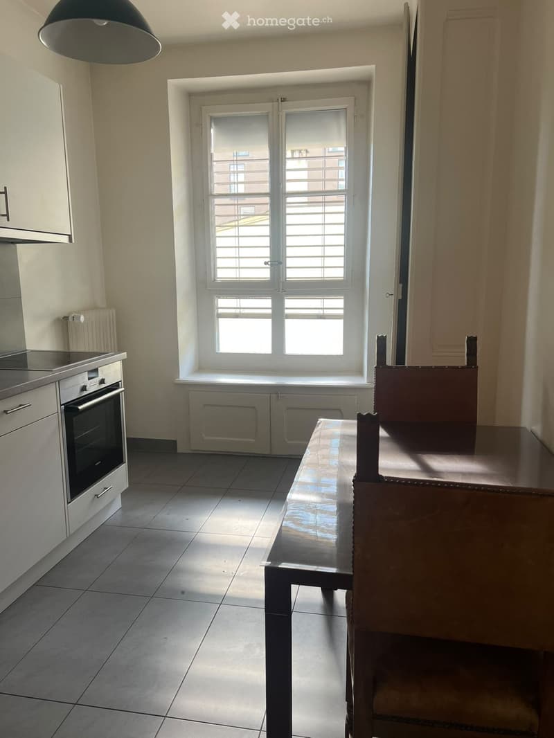 A modern kitchen with tiled flooring, white cabinets, an oven, and a dining table with chairs near a large window with blinds.