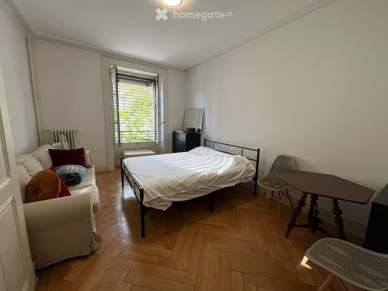 A small, furnished bedroom with a bed, sofa, small table, and chair. The room features wooden flooring and a window with blinds.