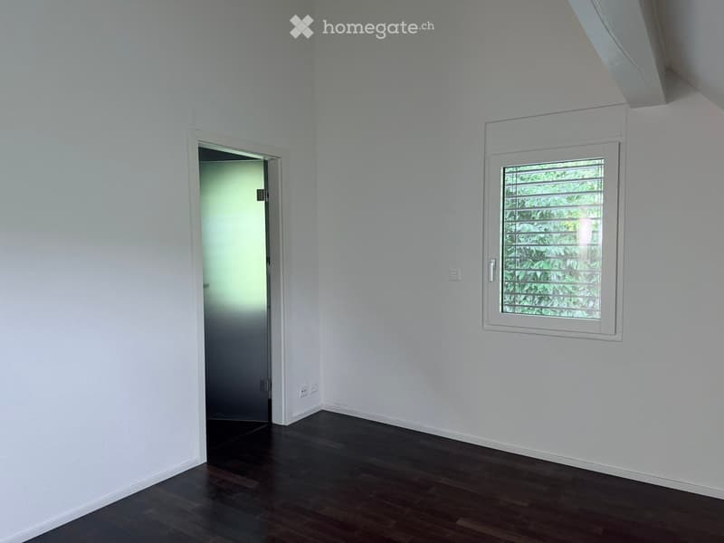 Empty room with white walls, dark wooden flooring, and a window with blinds. A door leads to another space.