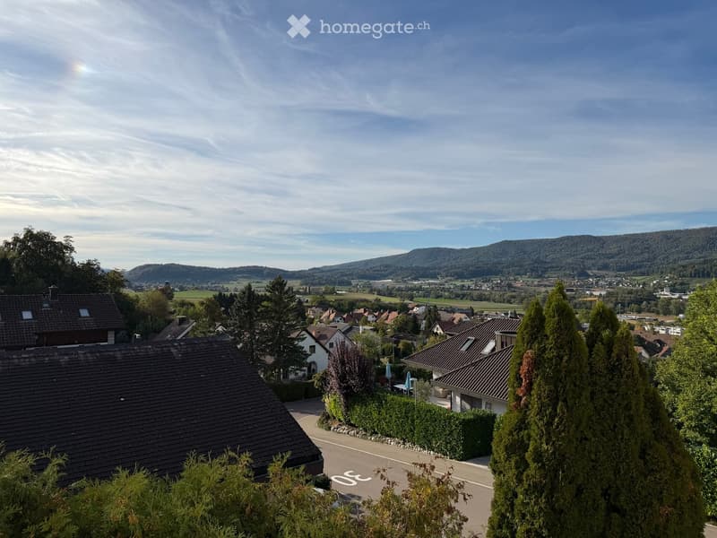 Residential area with multiple houses, surrounded by trees and greenery, with a scenic view of hills and countryside in the background.