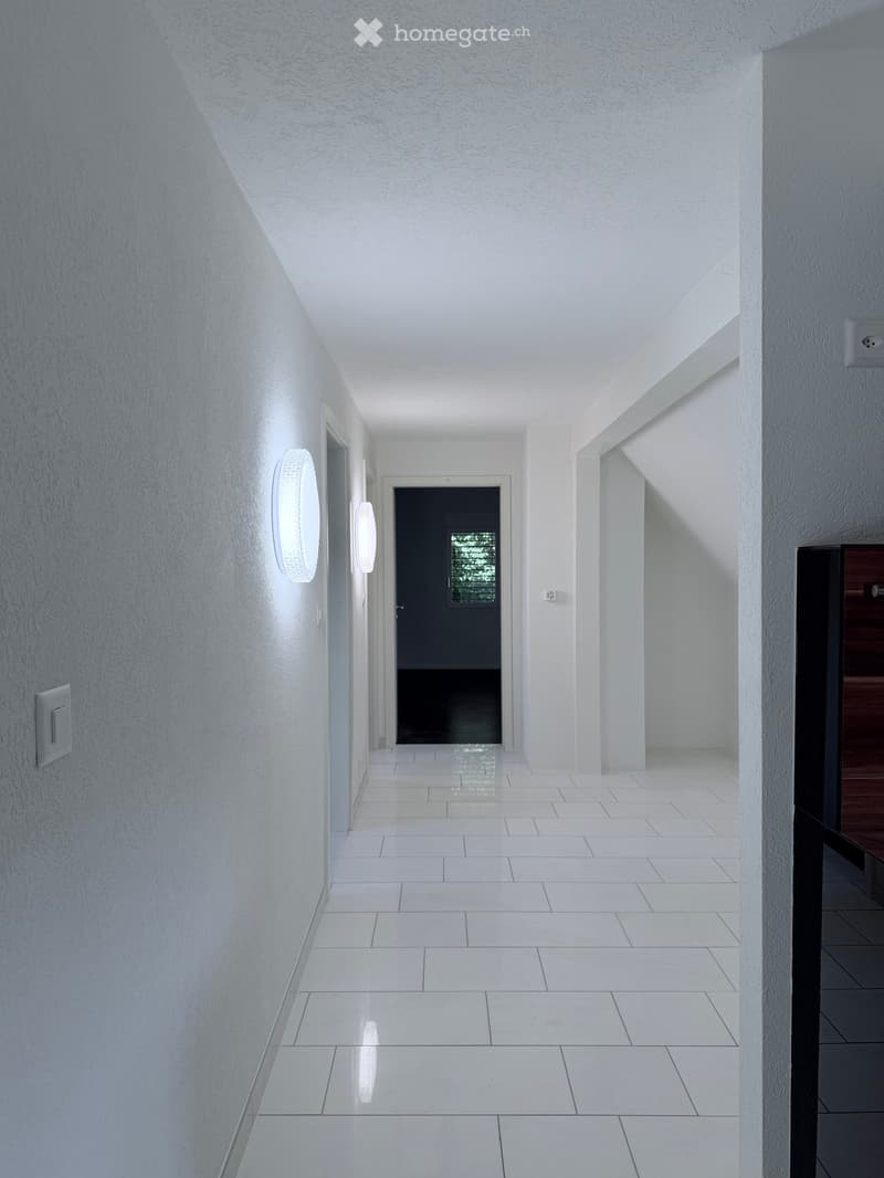 Modern hallway with white walls, white tiled floor, and recessed lighting. Features a black door at the end and a small wooden bench on the right side.