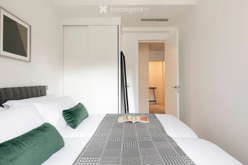 Modern bedroom with a neatly made bed, green throw pillows, a grey headboard, and a picture frame on the wall. An open door leads to another room with a stool visible.