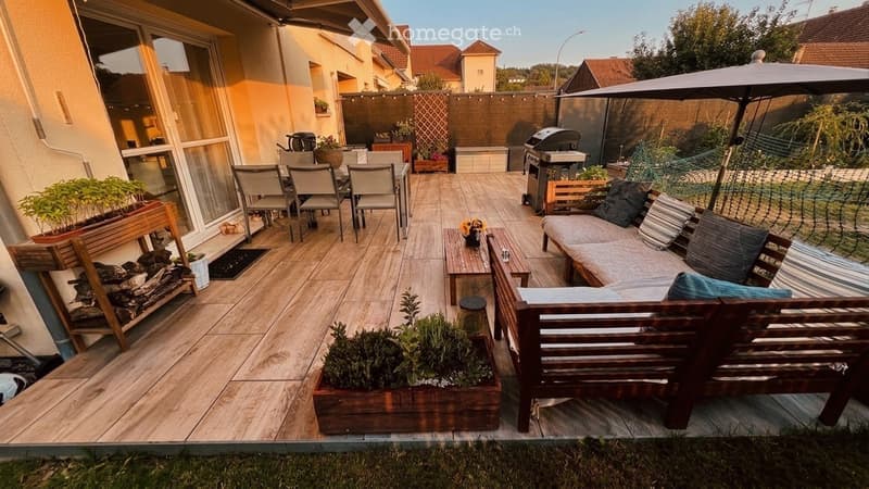 Wooden deck patio with outdoor furniture including a sofa, dining table, chairs, and a grill. Potted plants and a fire pit are also present.