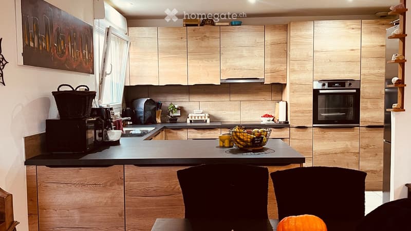 Modern kitchen with light wood cabinets, black countertop, and stainless steel appliances. Features include a coffee maker, fruit bowl, and a small table with chairs.