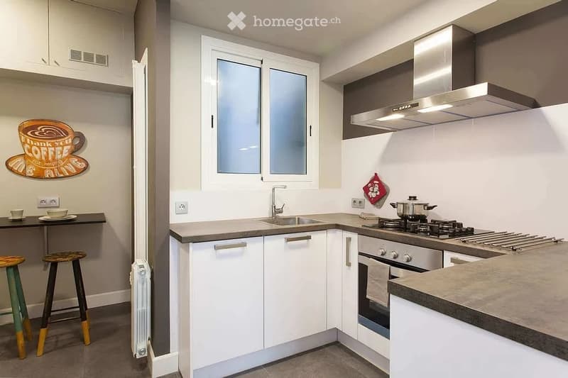 Modern kitchen with white cabinets, stainless steel appliances, a sink, stove, and oven. Includes a small table with two stools and a decorative wall sign.