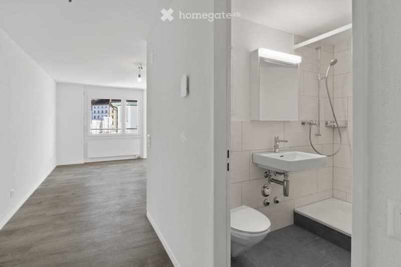 Modern bathroom with a toilet, sink, shower, and mirror. The space features light grey tiles, a wooden floor, and a window offering a view of the city.