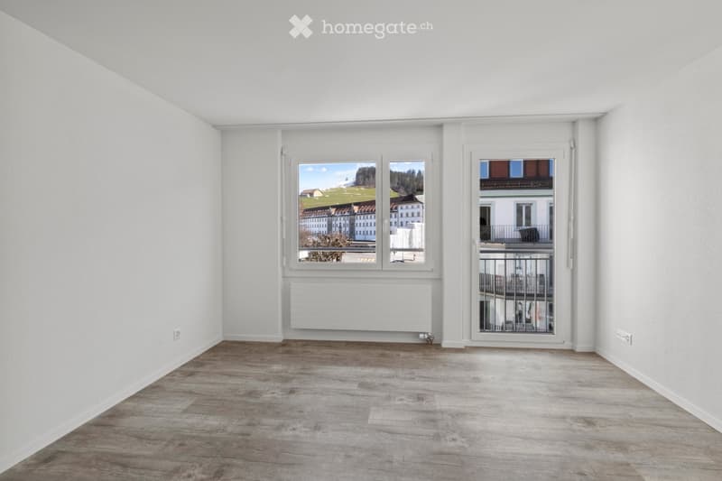 Empty room with wooden flooring, white walls, large windows, and a door leading to a balcony.