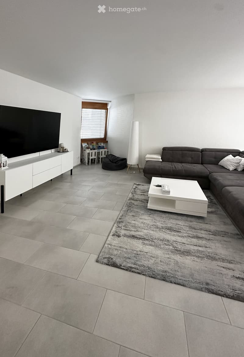 Modern living room with a grey sectional sofa, white coffee table, TV stand, floor lamp, and a small table with chairs near a window.