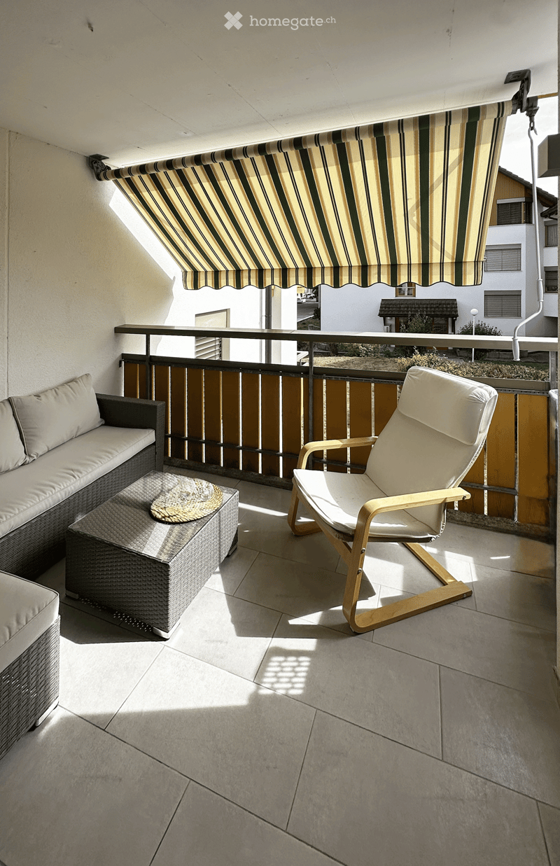 Balcony with a striped awning, a wicker sofa, a white wooden chair, and a small round table with a woven cushion.