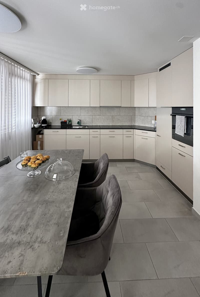 Modern kitchen with white cabinets, grey tiled backsplash, and a central island with a marble countertop. The island features a glass dome and a plate of muffins, with two grey chairs around it. A sink, oven, and various small appliances are visible along