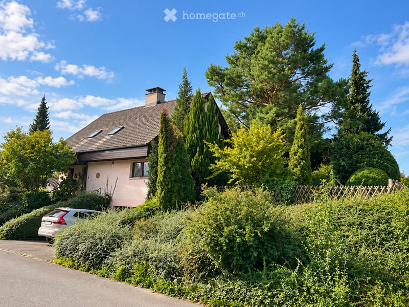 A two-story house with a tiled roof, surrounded by tall trees and greenery, with a parked car in the driveway.