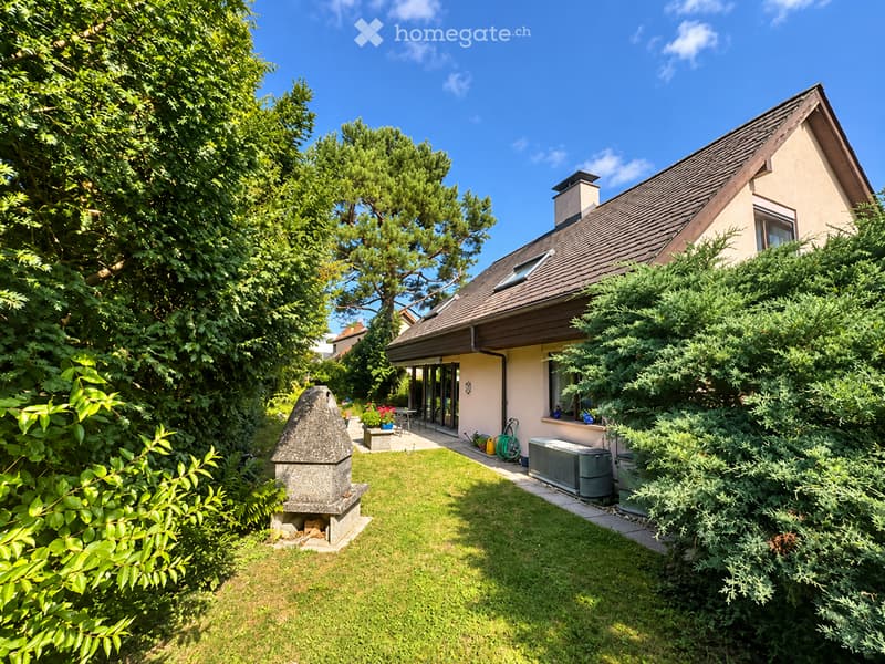 A well-maintained garden with a stone barbecue, neatly trimmed grass, and surrounding trees. The house features a modern design with large windows and a covered patio area.