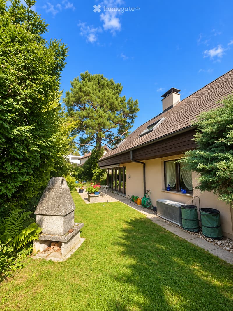 Well-maintained garden with a stone barbecue, green lawn, patio area with outdoor furniture, and a house with a tiled roof and large windows.