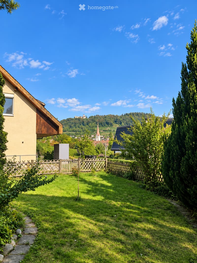 A well-maintained garden with a wooden fence, lush green grass, and trimmed bushes. The house has a light yellow exterior with a wooden roof section. The background features a scenic view of a town with a church tower and rolling hills.