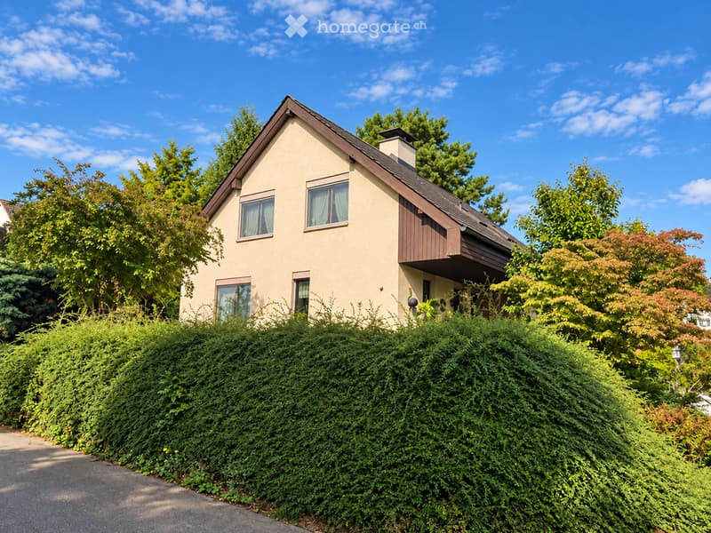 Single-family house with beige walls, dark brown roof, multiple windows, and a small balcony on the right side, surrounded by lush greenery and bushes.