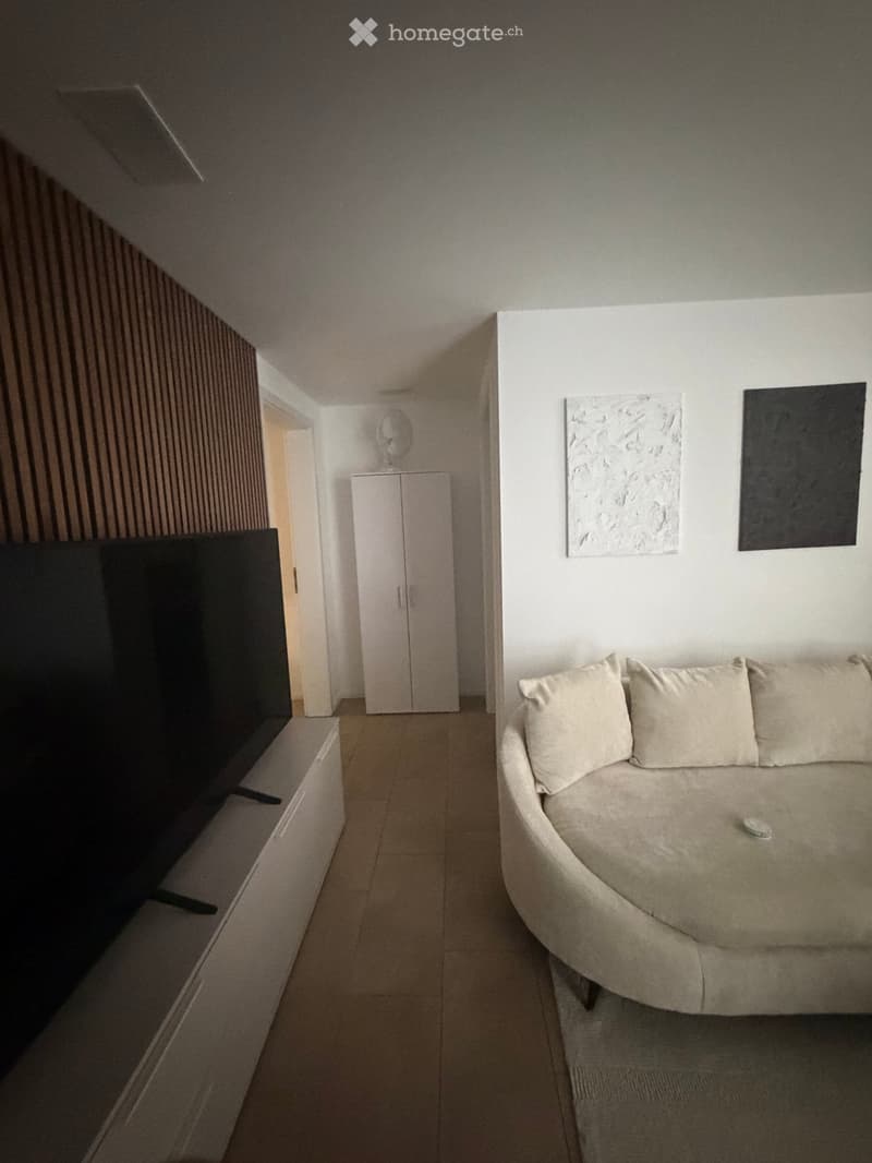Living room with a beige sofa, a white cabinet, a large screen on a stand, and a small table with a decorative item on top.