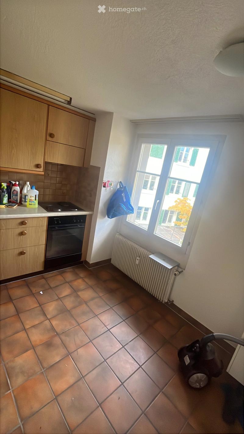A small kitchen with tiled flooring, wooden cabinets, an oven, a radiator, and cleaning supplies on the counter. A vacuum cleaner is placed on the floor near the window.