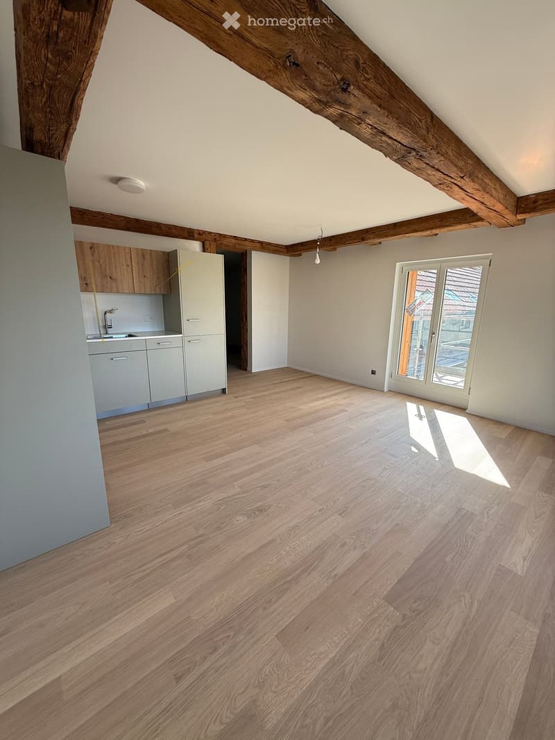 An empty attic room with wooden beams, light wooden flooring, and a small kitchenette area with cabinets and a sink.