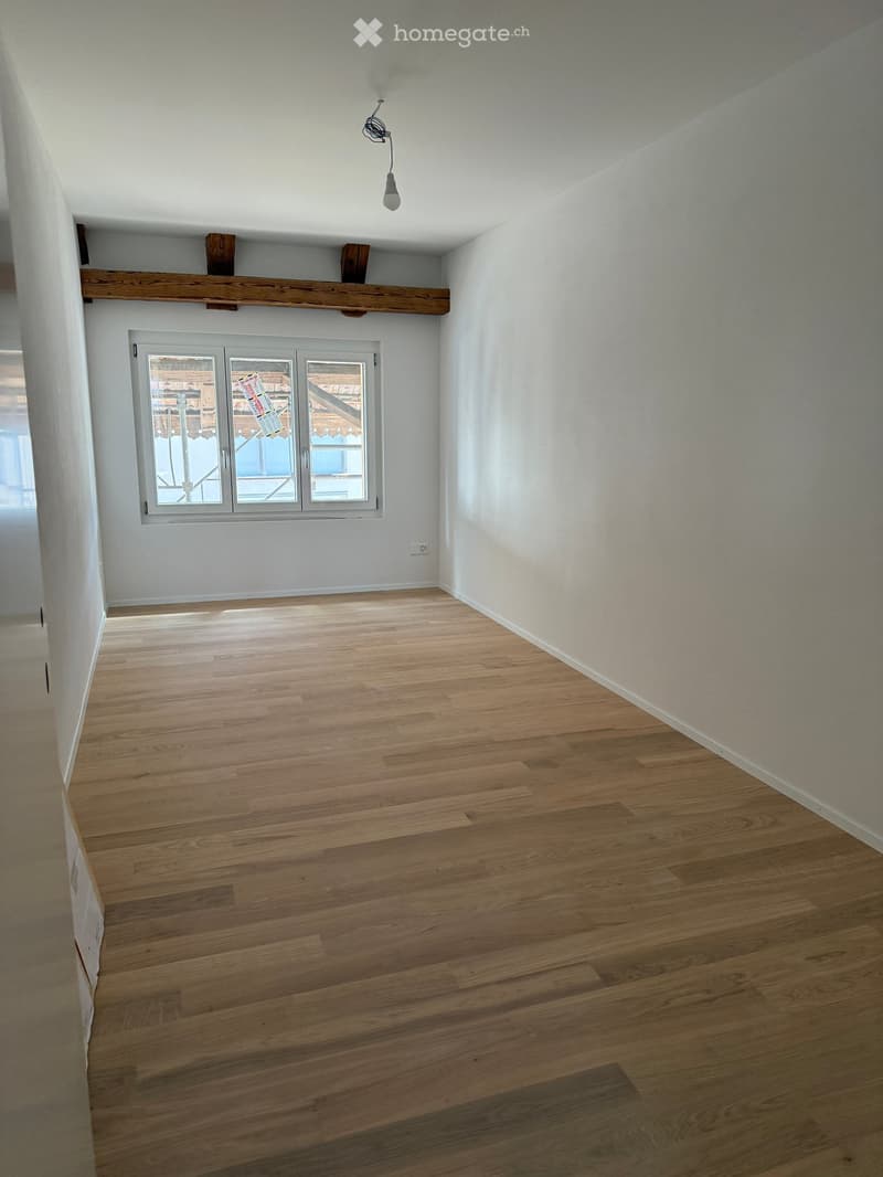 Empty room with wooden flooring, white walls, and a large window with a view of a wooden structure outside.
