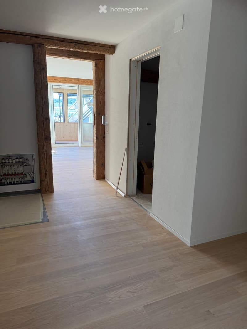 Empty room with wooden flooring, white walls, and exposed wooden beams. There is a doorway leading to another room and a visible heating system on the left side.