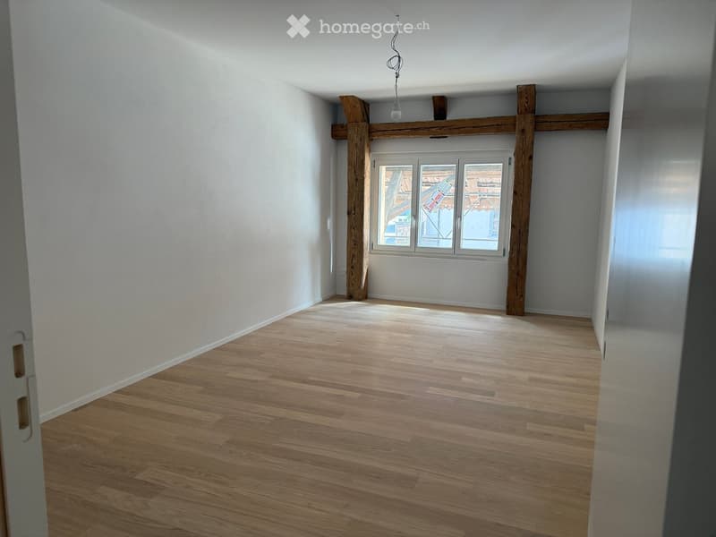 Empty room with wooden flooring, white walls, and exposed wooden beams. A single window with frosted glass panels is visible.