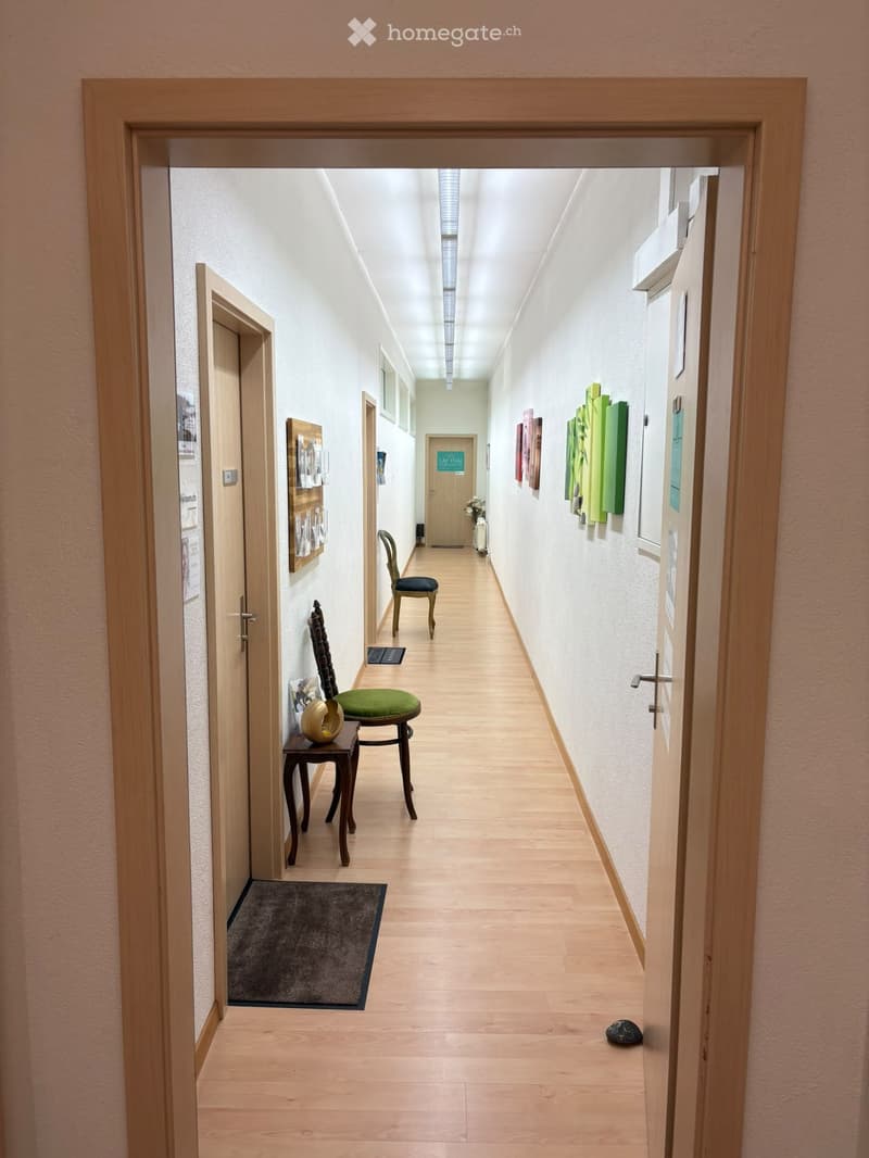 A long, well-lit hallway with light wooden flooring, white walls, and several doors. There are a few chairs and a small table with a decorative item placed along the walls.