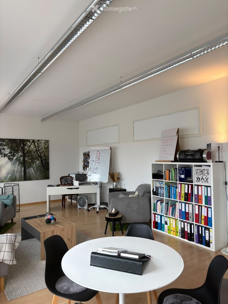 A modern, well-lit room with a mix of seating and work areas. Features include a grey sofa, a wooden coffee table, a white round table with chairs, a bookshelf filled with books and binders, a desk with a laptop, and a whiteboard with notes.