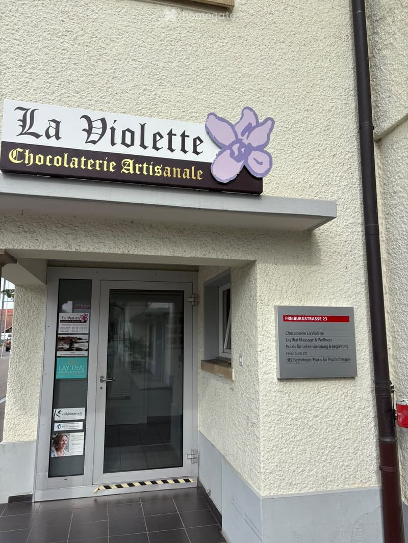Facade of a two-story building with a sign reading "La Violette Chocolaterie Artisanale" and a secondary sign indicating "FREIBURGSTRASSE 23" and listing services such as "Chocolaterie La Violette," "LayThai Massage & Wellness," and "Praxis für Lebensbera