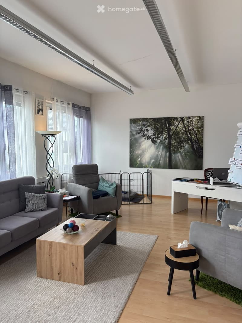 Modern living room with grey sofa, wooden coffee table, desk, and large window with curtains. Features a grey armchair, floor lamp, and artwork on the wall.