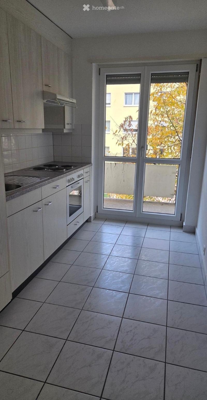 Empty kitchen with white tiled floor, white cabinets, and a large window with blinds overlooking a view of trees and a neighboring building.