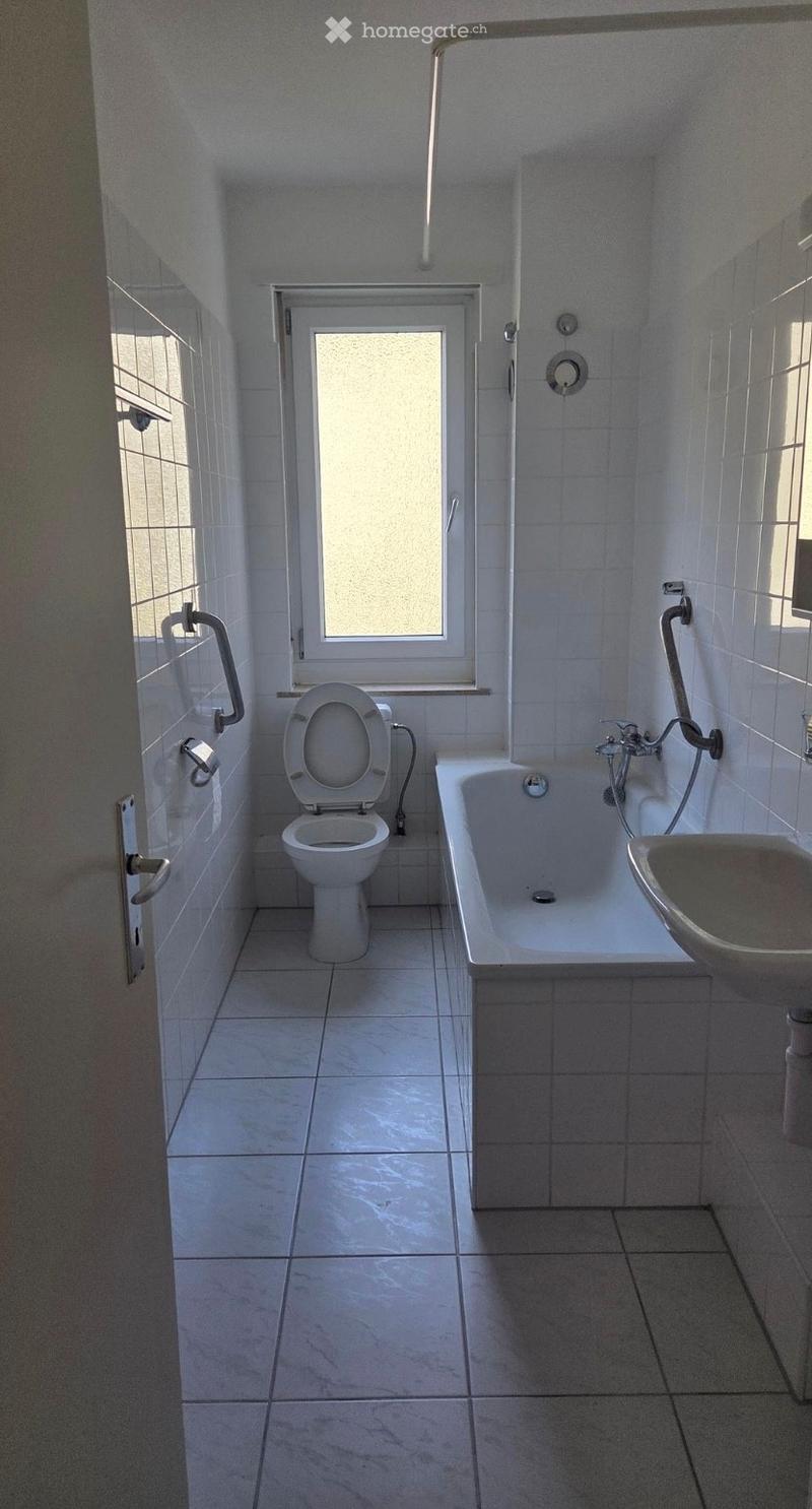 A small bathroom with white tiled walls and floor, featuring a toilet, bathtub, sink, and showerhead. The window has frosted glass, and there are grab bars on the walls for safety.