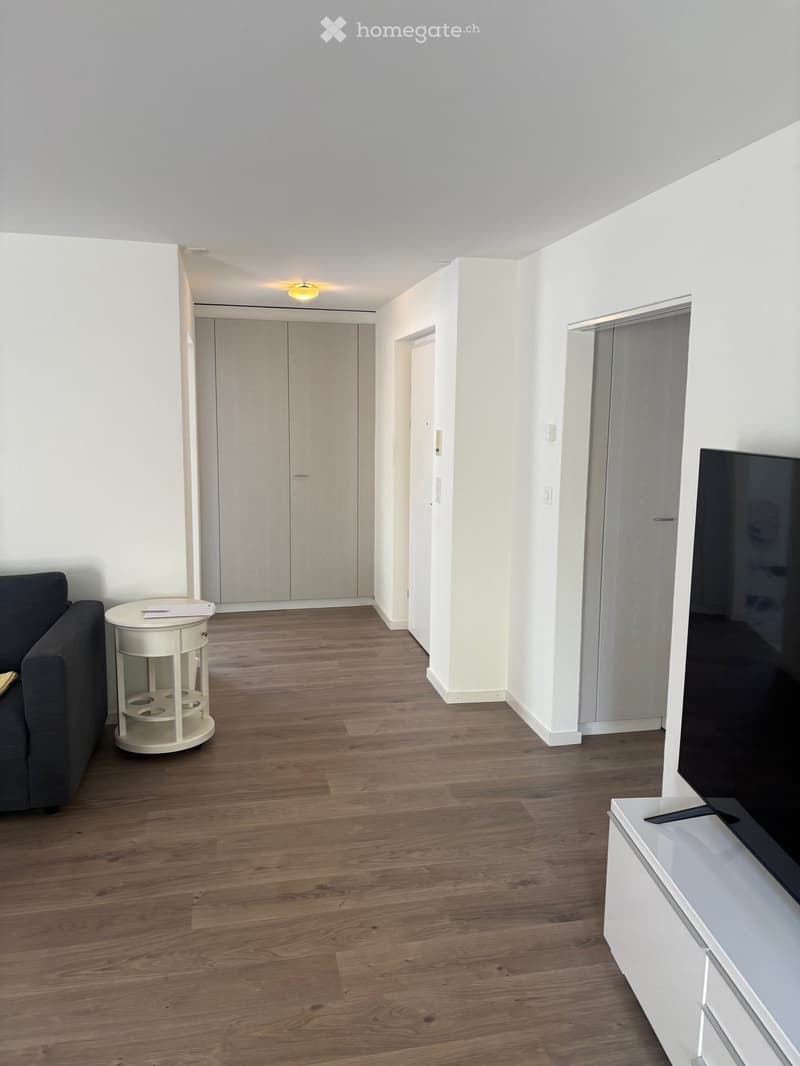 A modern living room with wooden flooring, white walls, and a grey sofa. A small white table is near the sofa, and a television is mounted on the wall.