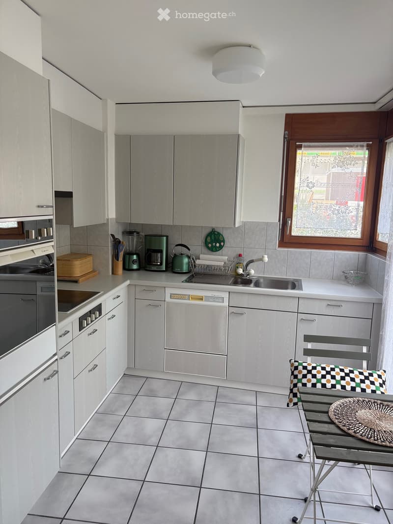 Modern kitchen with white cabinets, stainless steel appliances, tiled floor, and a small seating area with a stool and chair.
