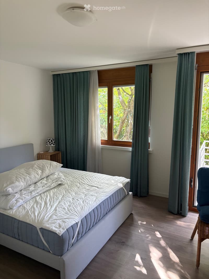 Modern bedroom with a large bed, nightstand, lamp, and a wicker chair near the window. The room features wooden flooring and large windows with green curtains.