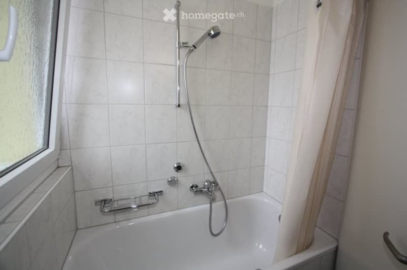 Bathtub with a handheld showerhead and a handrail for support, tiled walls, and a window with a curtain.